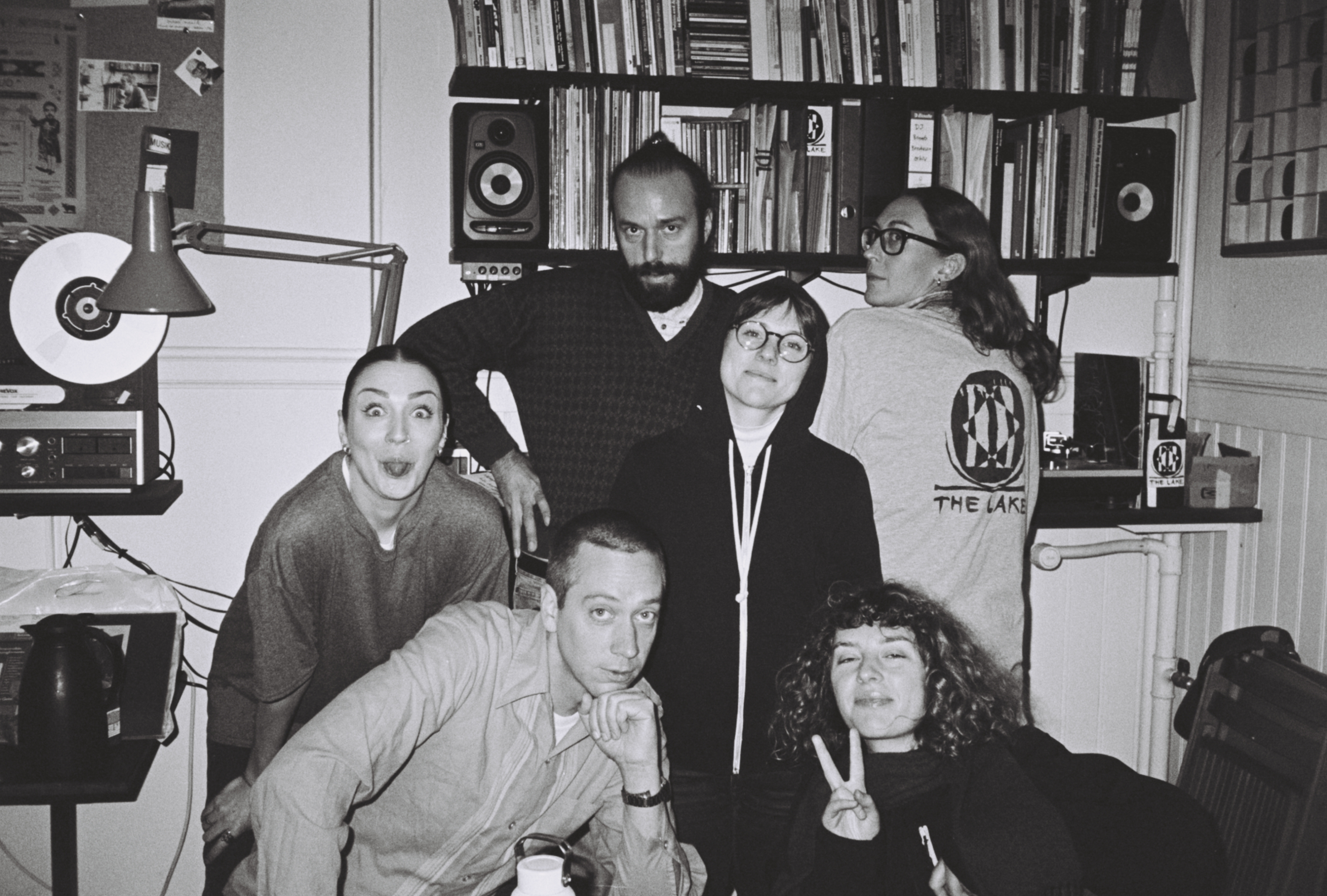 A black and white photo of six people posing while all looking into the camera. In the background there is sound equipment, speakers, and shelves full of CDs, vinyl and books.